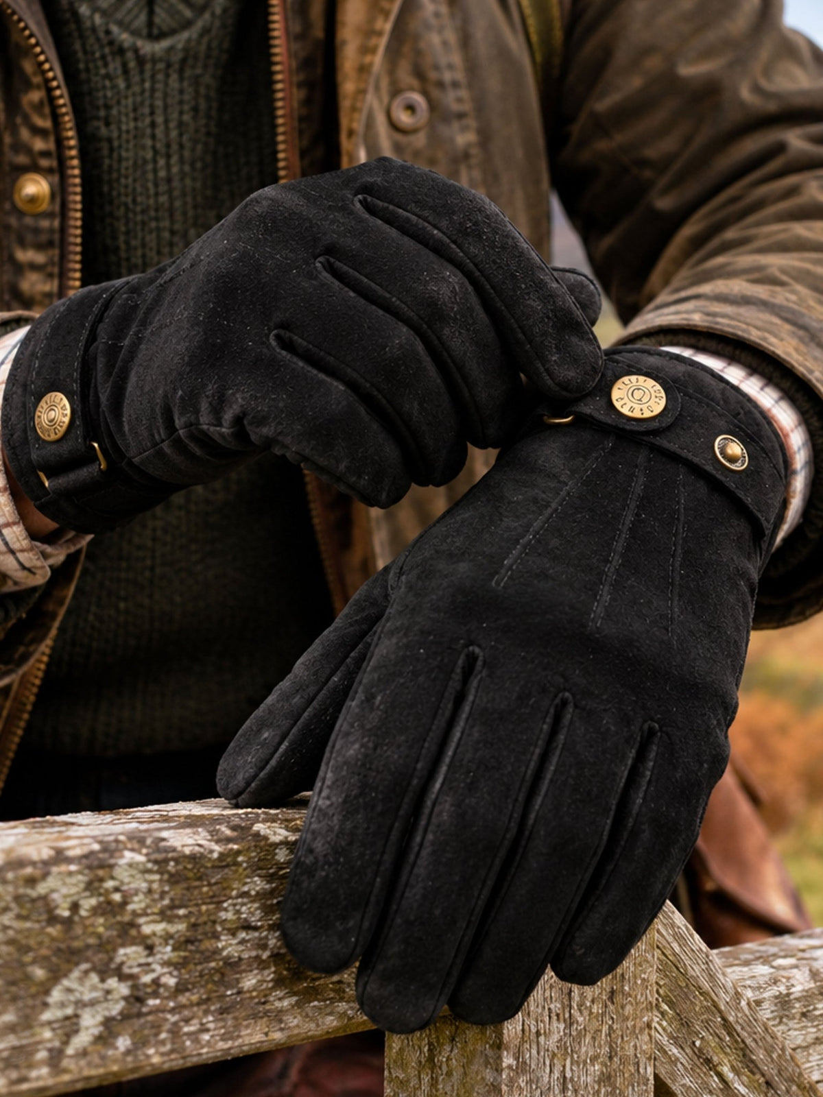 Dents men's black touchscreen suede gloves with button strap, worn with waxed jacket on a stone wall