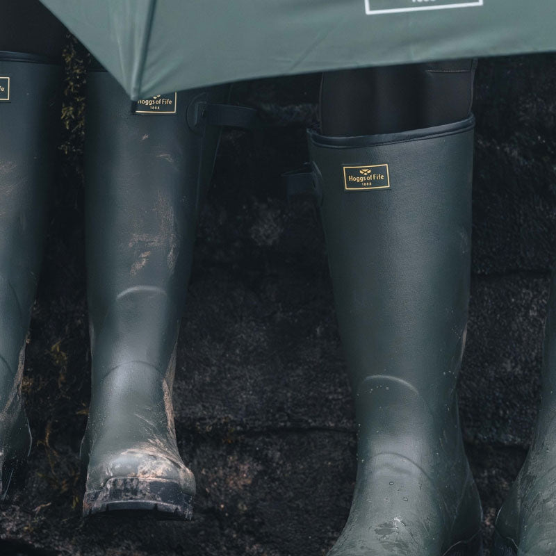 Hoggs of fife wellies - Four dark green rubber rain boots with gold-toned metal logo plates, worn by people sitting on a dark stone ledge outdoors.