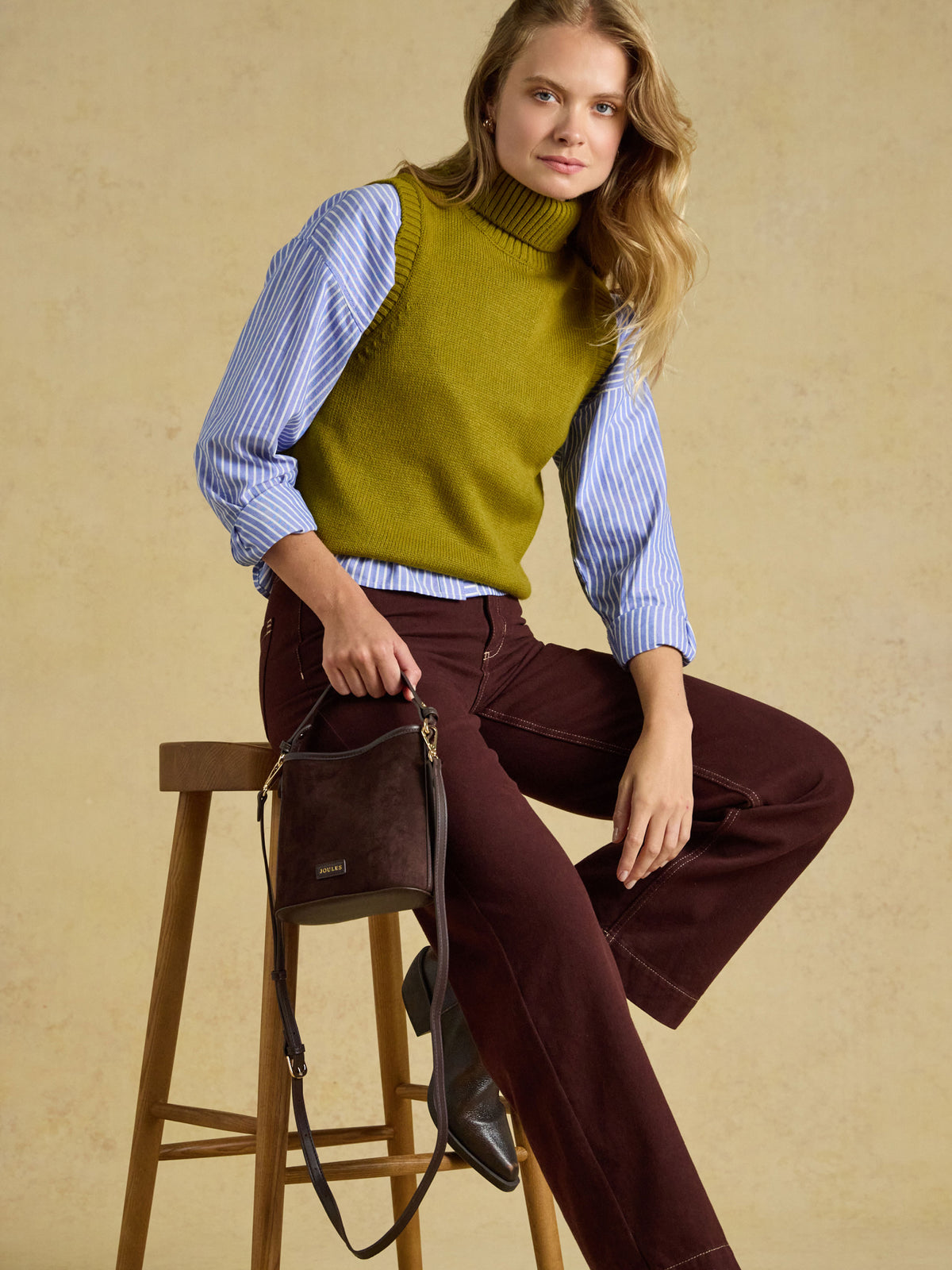 A woman models a knit turtleneck sweater vest in olive green, layered over a light blue and white striped long-sleeve shirt, paired with dark burgundy wide-leg pants and holding a dark brown leather shoulder bag while sitting on a wooden stool against a textured beige background.