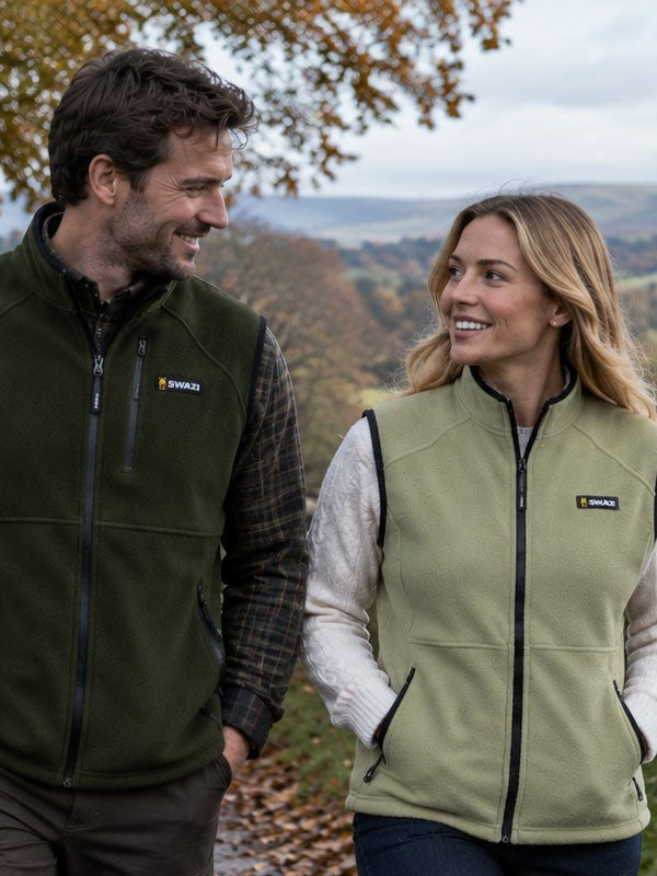 A man and a woman standing outdoors on a country path, both wearing Patagonia fleece vests. The man's vest is dark olive green, and the woman's vest is light sage green, with a scenic rural landscape with hills, trees, and a stone wall in the background.