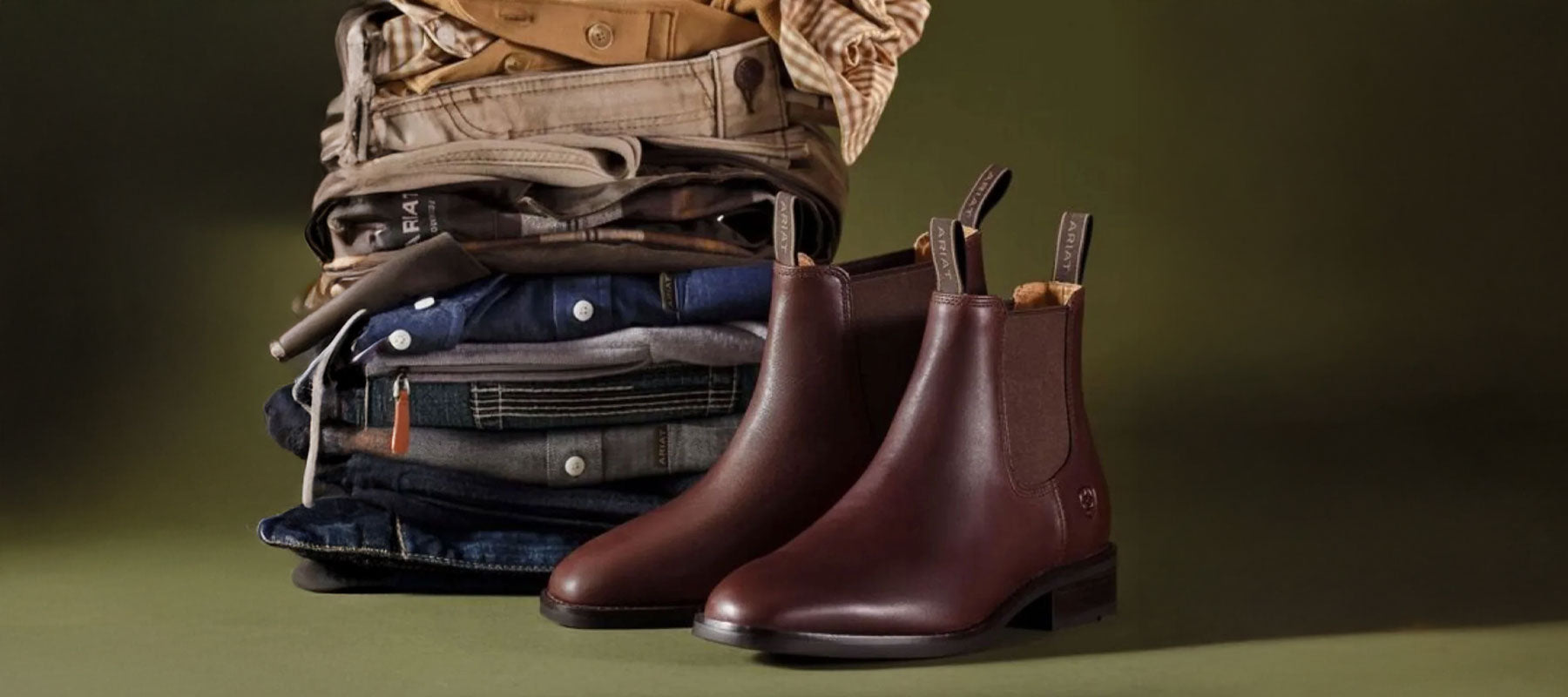A pair of dark brown leather Chelsea boots displayed in front of a stack of folded clothing, including jeans and a tan jacket, on a dark olive green background.