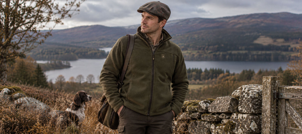 A man wearing an olive green fleece jacket, brown trousers, and a brown flat cap stands outdoors in a scenic landscape with a lake and mountains in the background. A dog is visible on the left side of the frame.