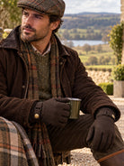 Man wearing Dents brown waxed cotton gloves with a waxed jacket and tartan scarf, holding a mug by a lake