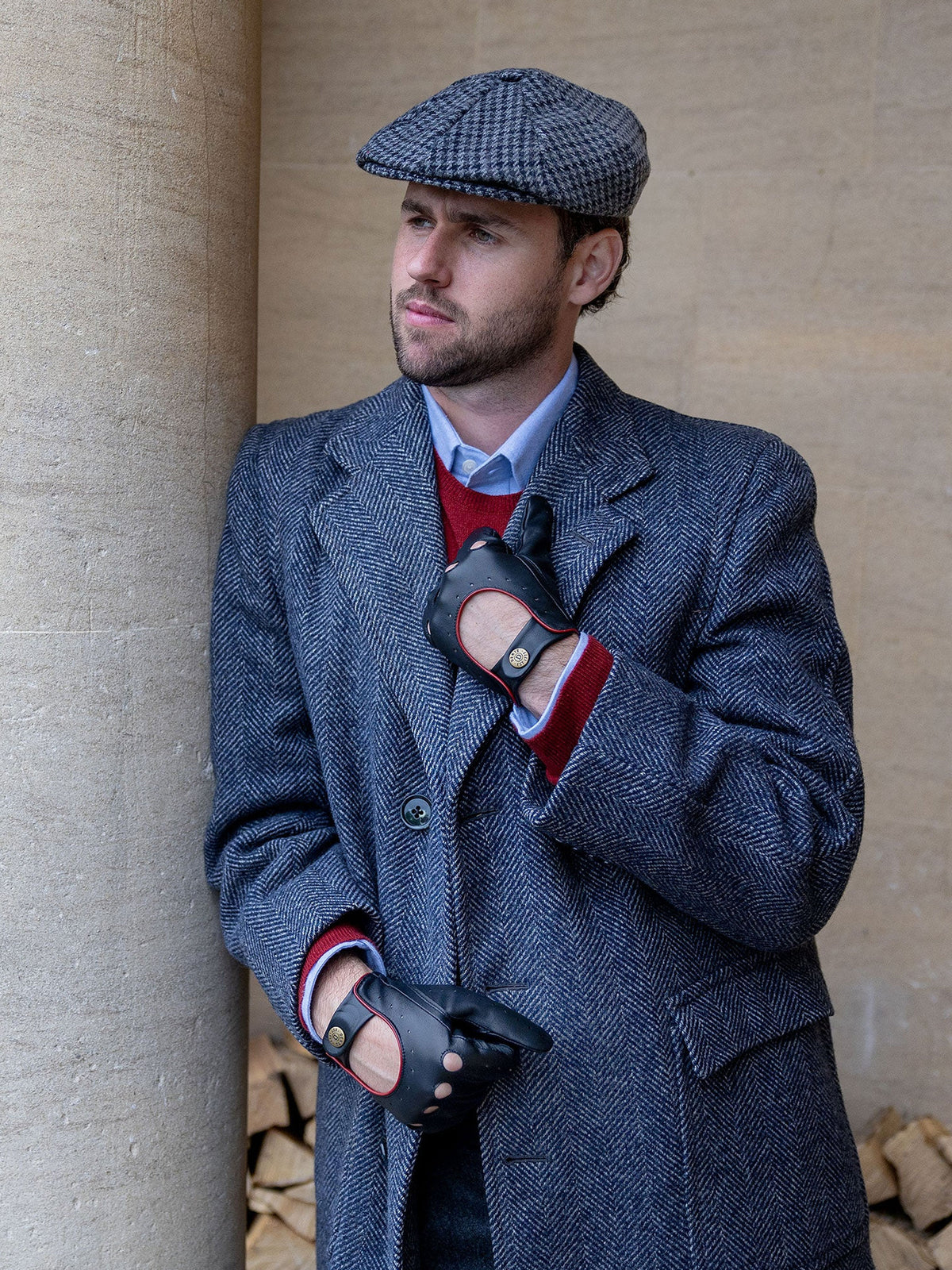 Man wearing Dents Delta black leather driving gloves with a blue herringbone coat, tweed flat cap and red jumper