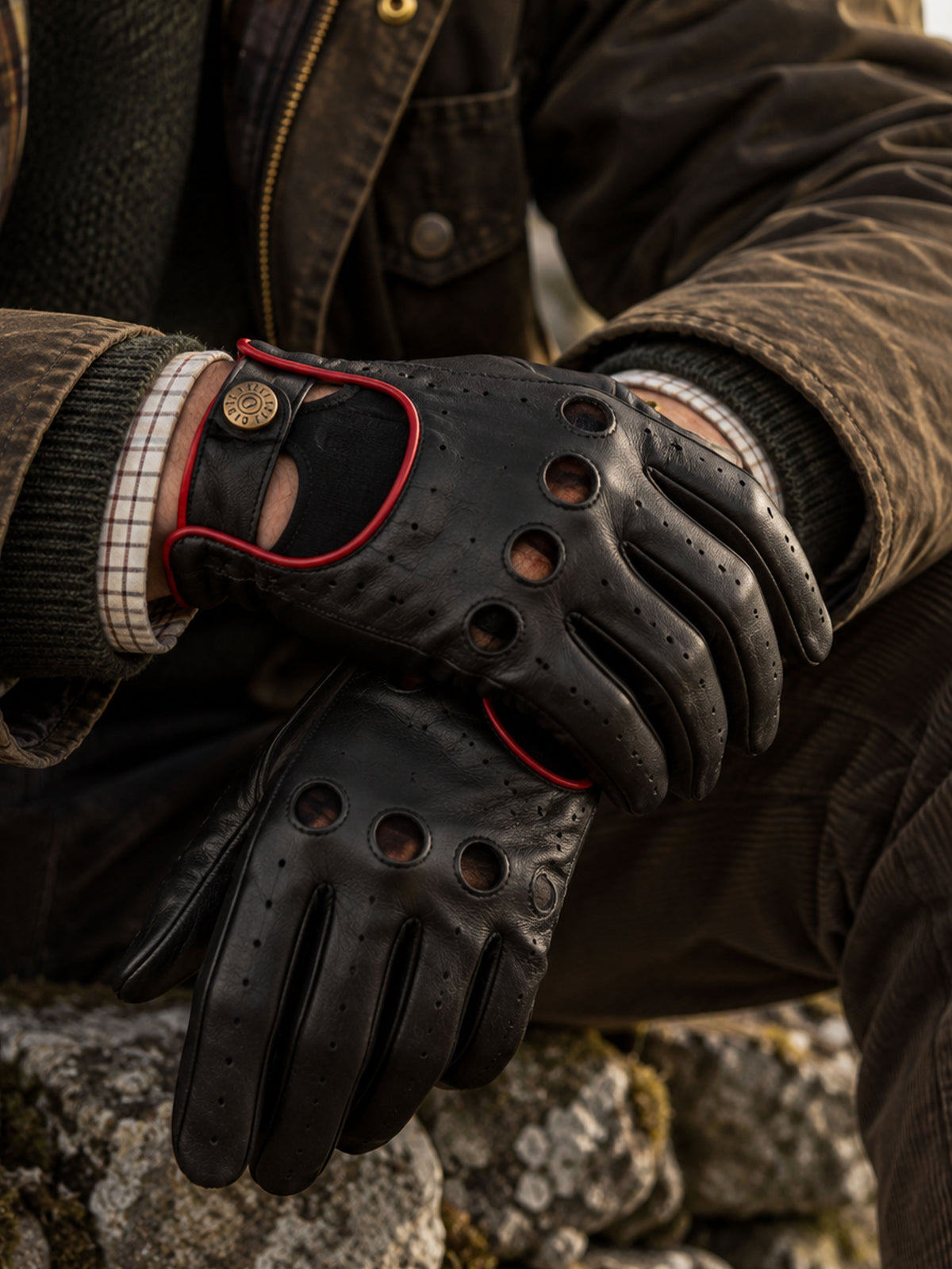 Dents Delta men's black leather driving gloves with red trim, worn with waxed jacket on a stone wall