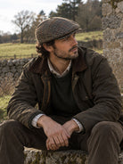 Man wearing a Dents glen plaid check brown tweed flat cap with a green waxed jacket, seated outdoors in the countryside