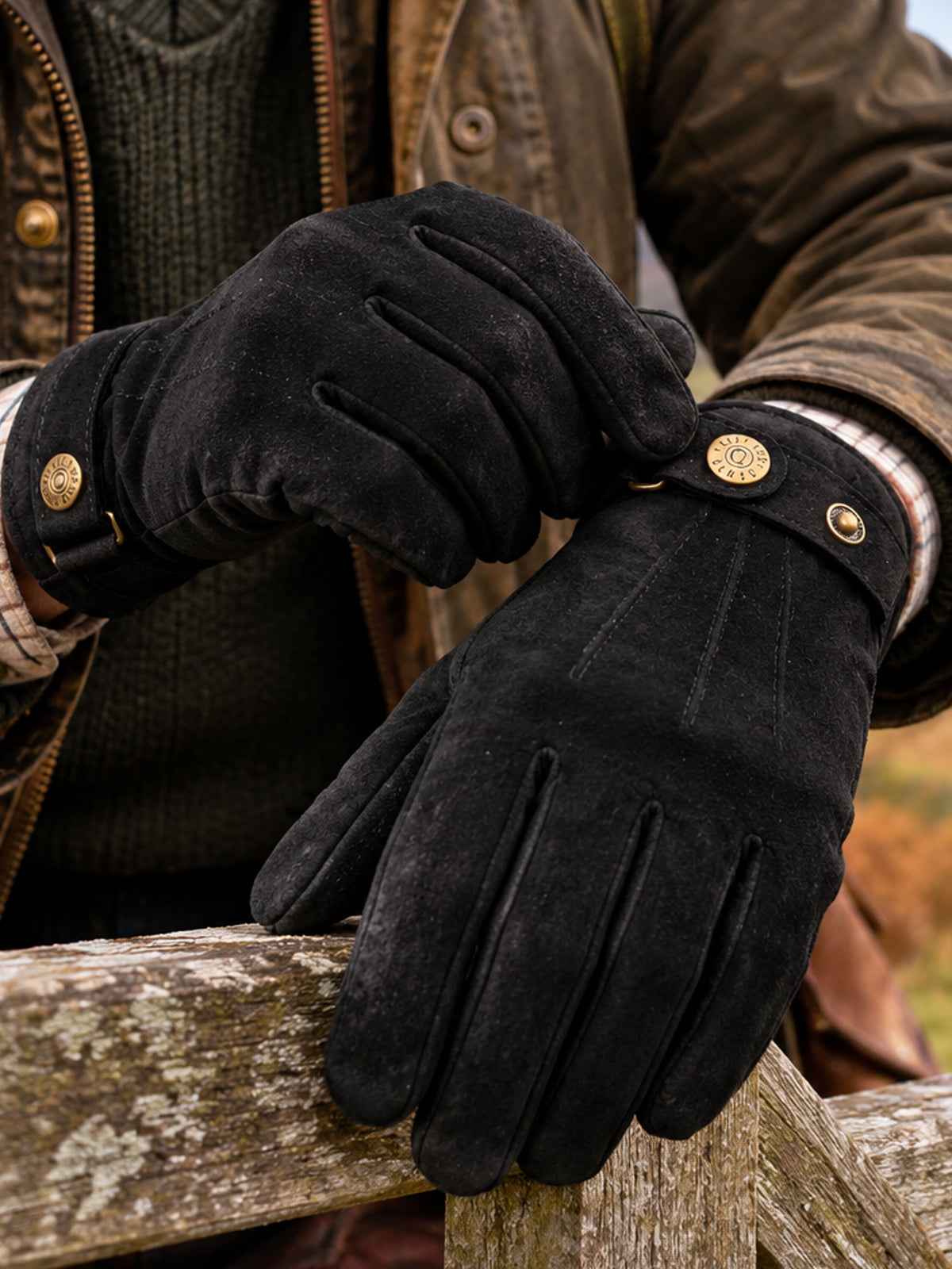 Dents men's black touchscreen suede gloves with button strap, worn with waxed jacket on a stone wall
