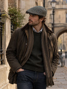 Dents Thirlmere men's olive herringbone tweed flat cap, worn with waxed jacket, scarf and jeans on a cobbled street