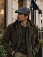 Dents Thirlmere men's charcoal herringbone tweed flat cap, worn with waxed jacket and scarf, front three-quarter view
