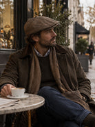 Dents Thirlmere men's chocolate brown herringbone tweed flat cap, worn with waxed jacket and scarf at a café table