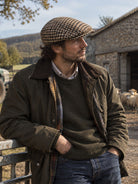 Dents Blashford men's brown dogtooth tweed flat cap, worn with waxed jacket outside a stone barn