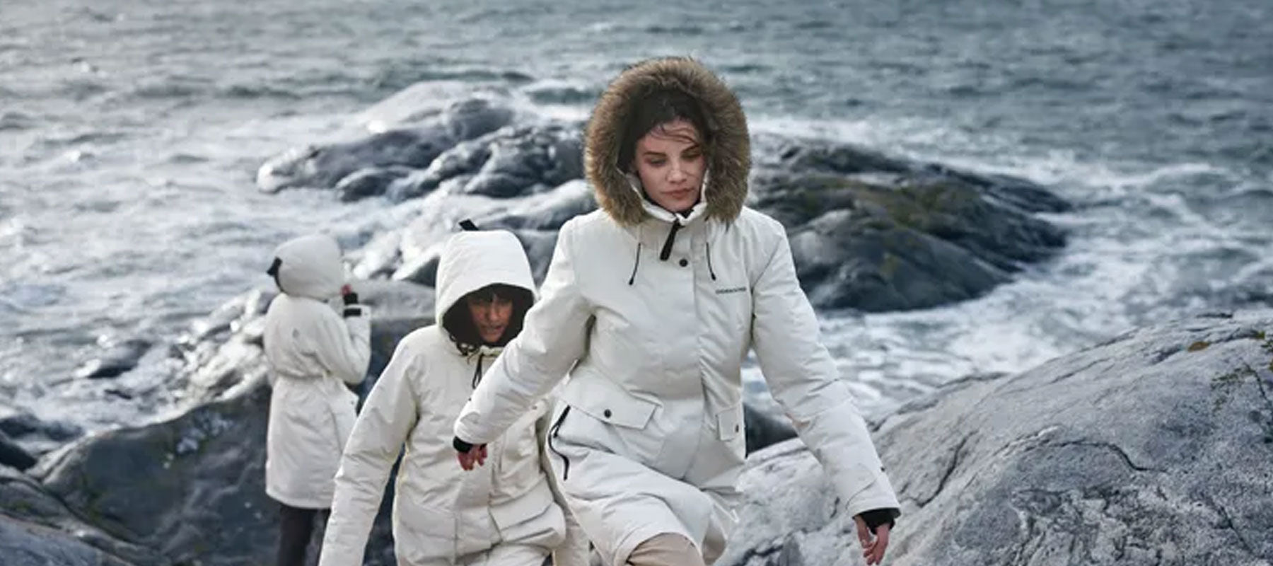 Woman in a white Didriksons parka with fur-trim hood on a rocky coast