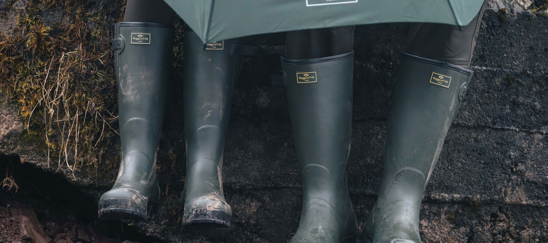 Four dark green rubber rain boots with gold-toned metal logo plates, worn by people sitting on a dark stone ledge outdoors.