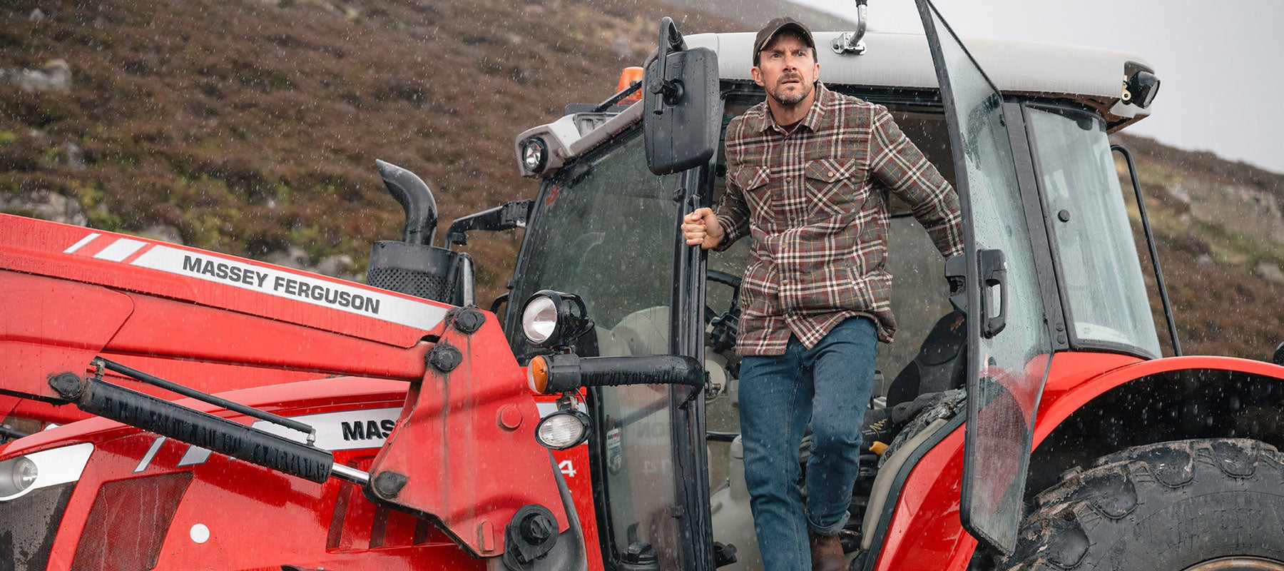 A man wearing a brown and beige plaid shirt and blue jeans stands on the open door of a red Massey Ferguson tractor in an outdoor field.