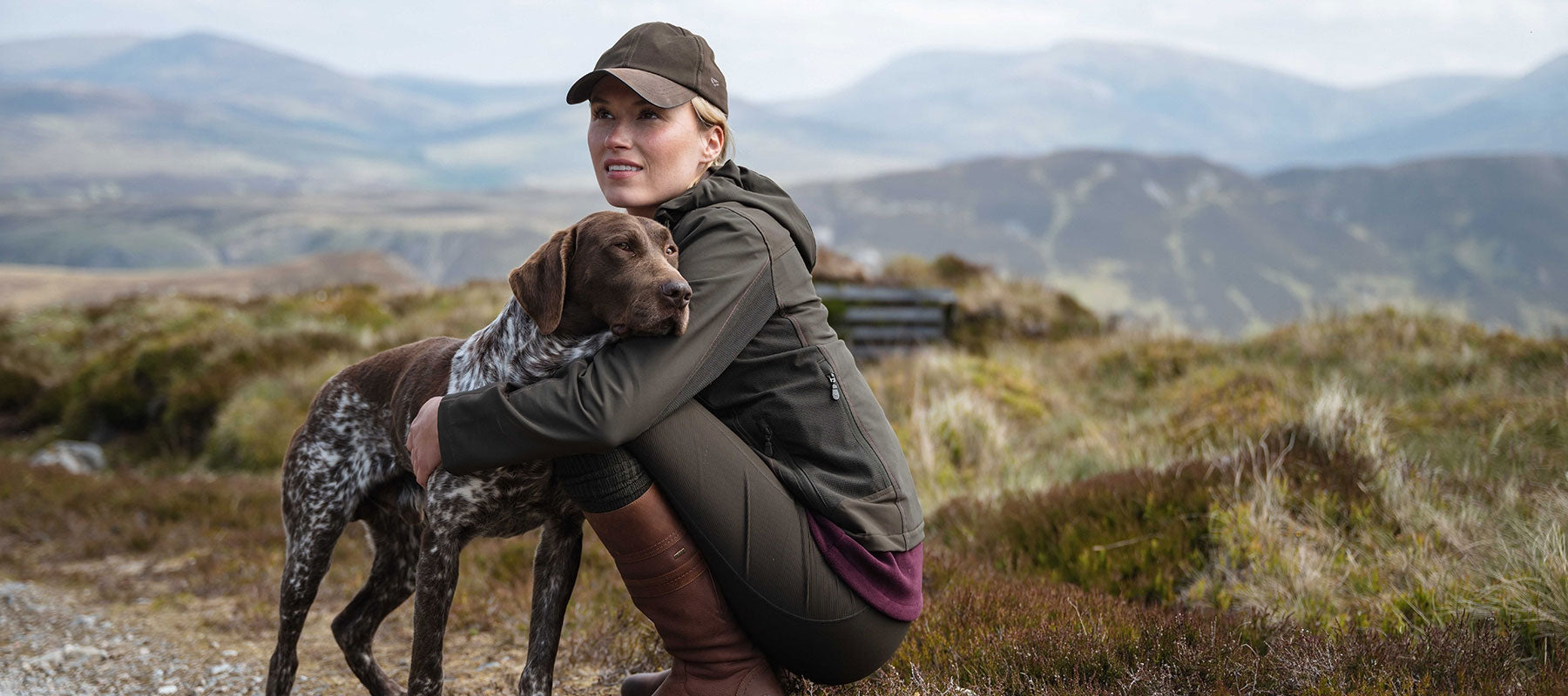 A woman crouches outdoors in a mountain landscape, embracing a brown and white spotted dog. She wears a dark olive jacket, dark leggings, a brown cap, and tall brown boots.