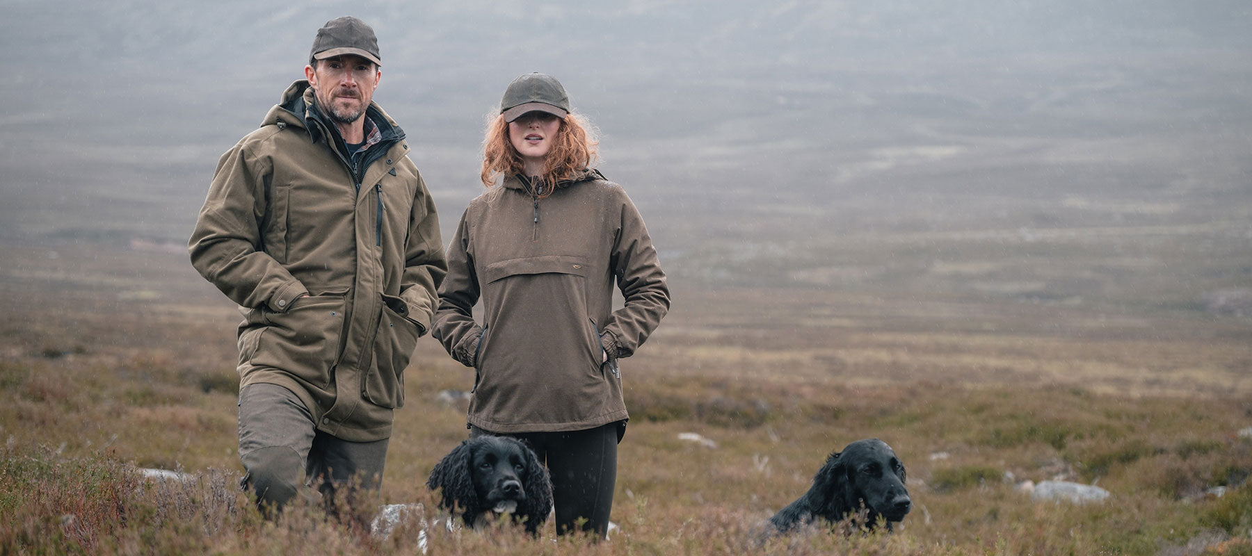 A man and a woman wearing matching olive green outdoor jackets and caps stand in a misty, grassy landscape with two black dogs in the foreground.