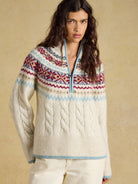 A woman wearing a cream cable-knit half-zip sweater with a multicolor Fair Isle yoke pattern in red, blue, and white, paired with cream pants, set against a warm beige textured background.