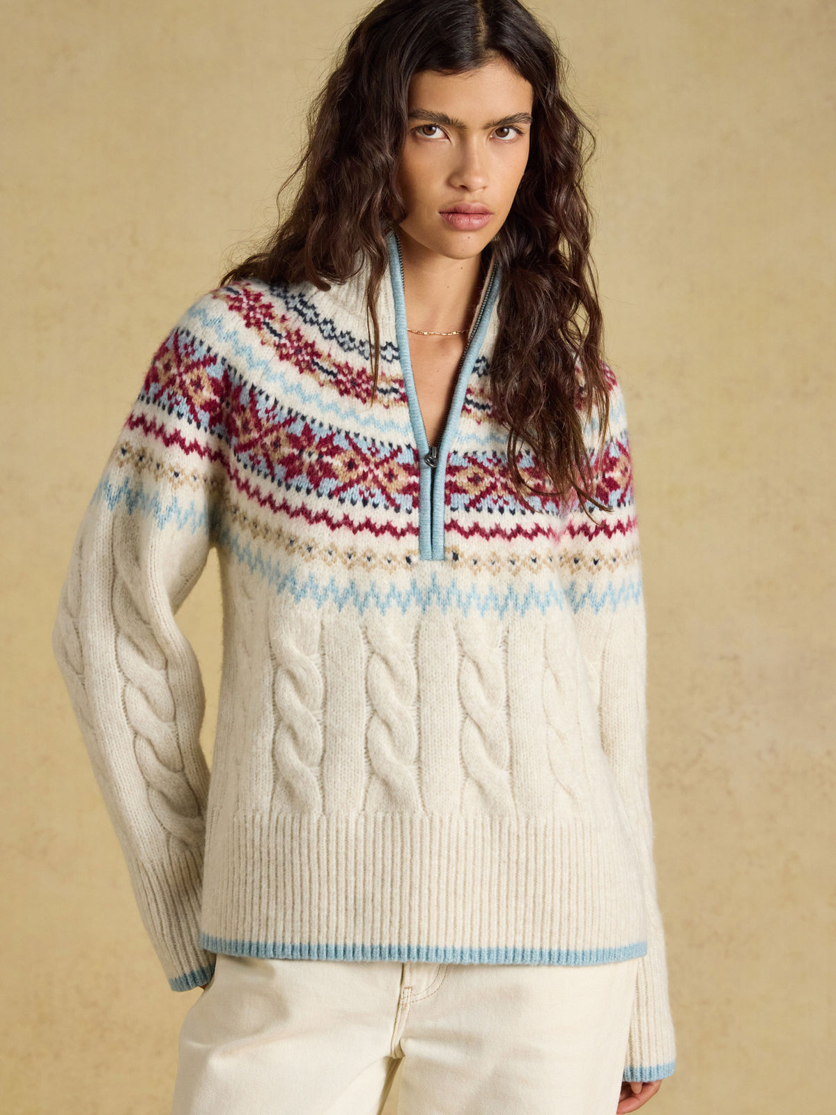 A woman wearing a cream cable-knit half-zip sweater with a multicolor Fair Isle yoke pattern in red, blue, and white, paired with cream pants, set against a warm beige textured background.