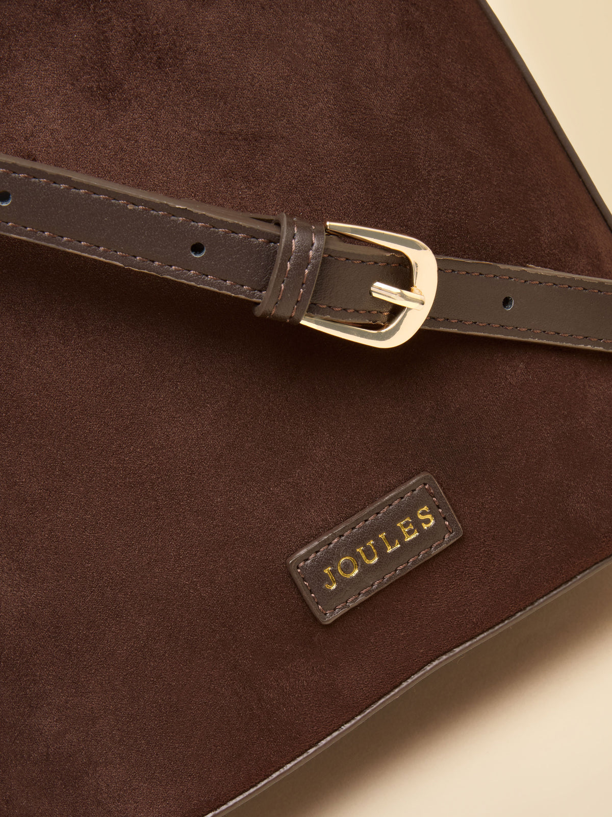 Close-up of a dark brown suede shoulder bag with a matching brown leather strap and gold-tone buckle, featuring a small gold JOULES brand patch.