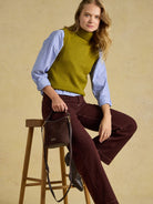 A woman models a knit turtleneck sweater vest in olive green, layered over a light blue and white striped long-sleeve shirt, paired with dark burgundy wide-leg pants and holding a dark brown leather shoulder bag while sitting on a wooden stool against a textured beige background.