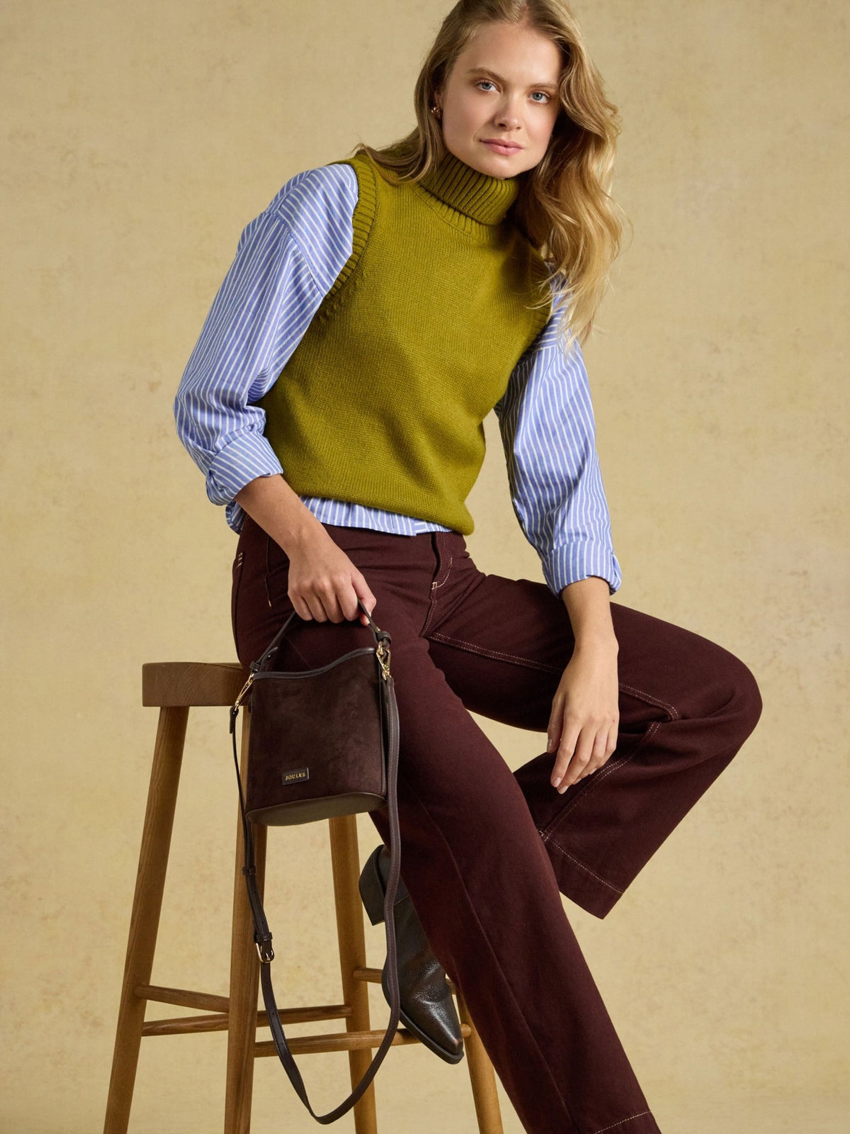 A woman models a knit turtleneck sweater vest in olive green, layered over a light blue and white striped long-sleeve shirt, paired with dark burgundy wide-leg pants and holding a dark brown leather shoulder bag while sitting on a wooden stool against a textured beige background.