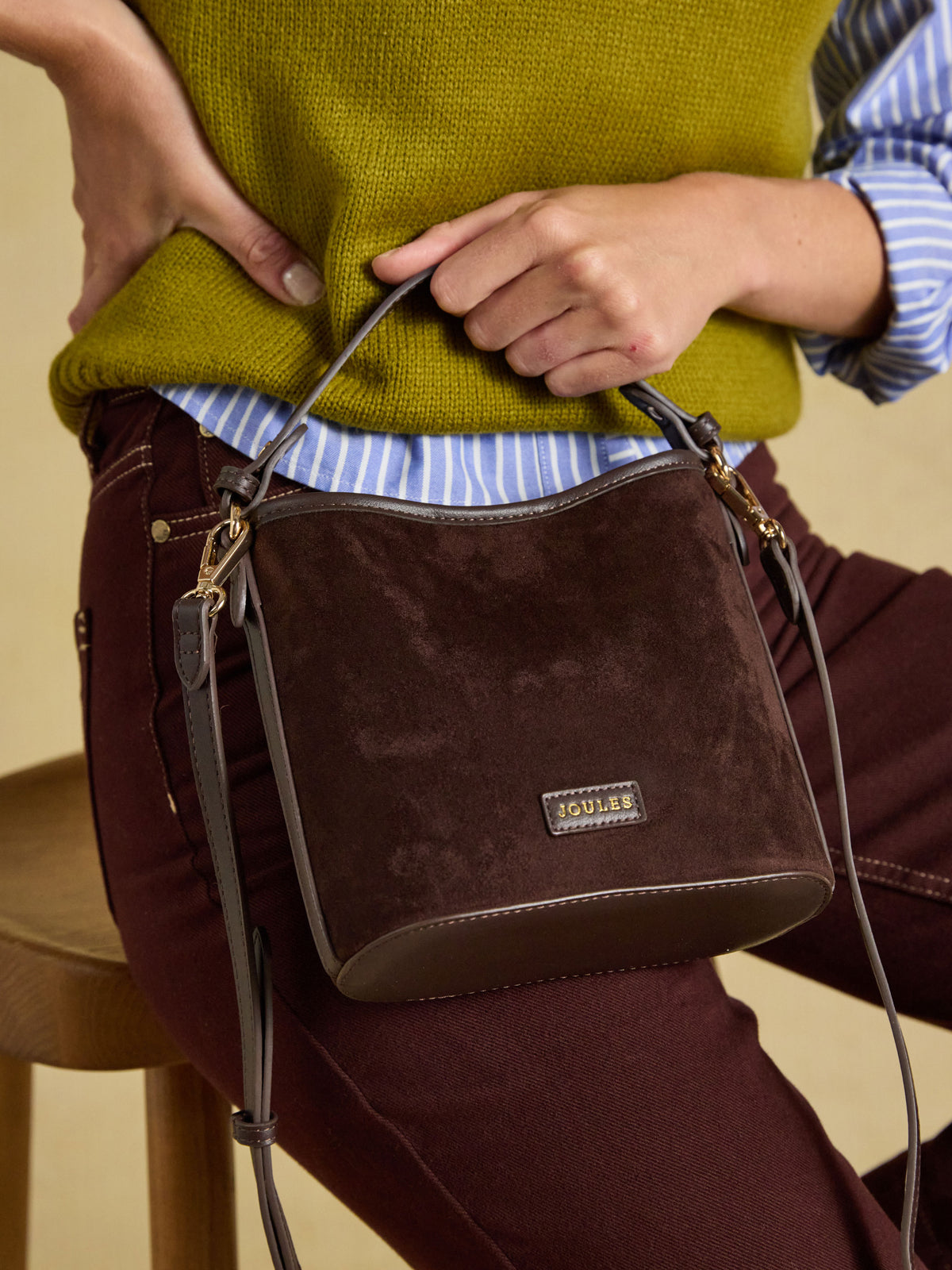 A person wearing a mustard green sweater, blue and white striped shirt, and burgundy pants carries a dark brown suede crossbody bag with a top handle and a small Jowell brand plaque on the front.