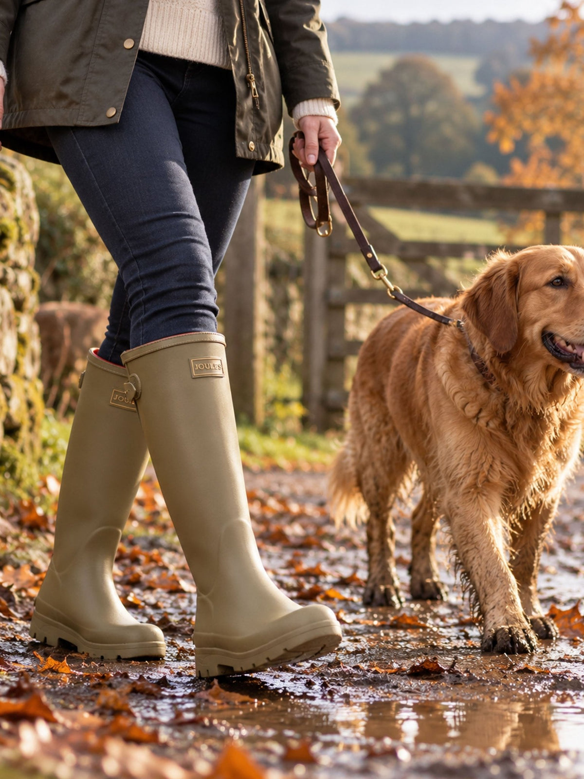 Woman walking a golden retriever on a muddy country path wearing Joules Luxe khaki green neoprene lined tall wellies