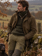 Man wearing a Seeland Apex One Hundred merino neck gaiter in Pine Green with a waxed jacket, leaning on a stone wall with a spaniel