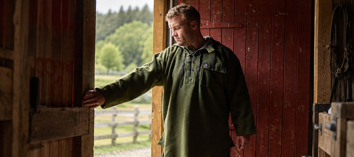 A man wearing an olive green long-sleeve henley shirt stands in a rustic barn doorway, looking off-camera. The background shows a red wooden door, wooden beams, and a blurred outdoor landscape with green trees.