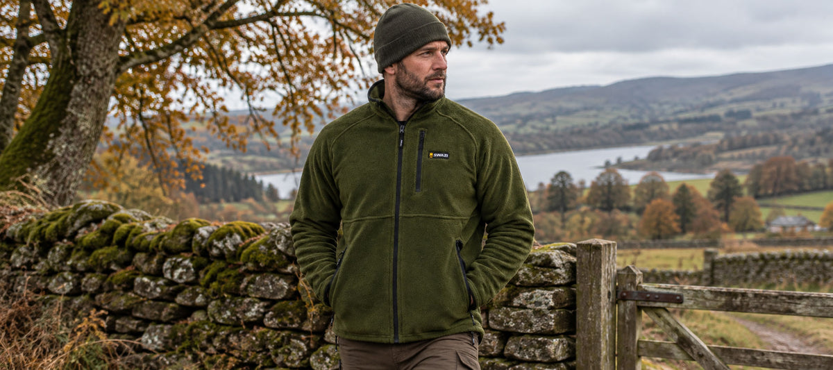 A man wearing an olive green fleece jacket and a dark beanie stands outdoors in front of a stone wall, with a lake, hills, and autumn trees in the background. A small yellow and blue Patagonia logo is visible on the left chest of the jacket.