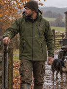 A man wearing an olive green Patagonia hooded jacket, brown beanie, and muddy trousers stands outdoors next to a wooden gate, with a dog in the background on a farm path.