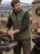 A man wearing an olive green Patagonia zip-up puffer vest over a dark long-sleeve shirt stands outdoors near a stone structure and fire pit, packing a brown bag on a table. A black dog is visible at the lower right, with a mountain landscape in the background.