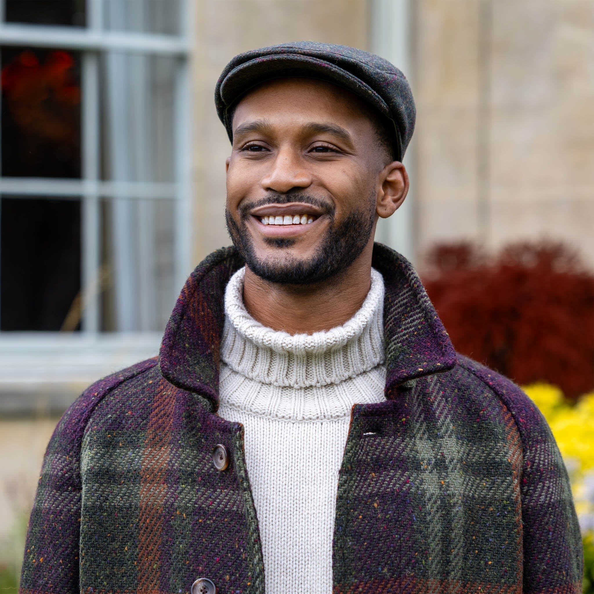 Dents men's olive windowpane check tweed flat cap, front view worn with plaid coat and cream roll neck jumper