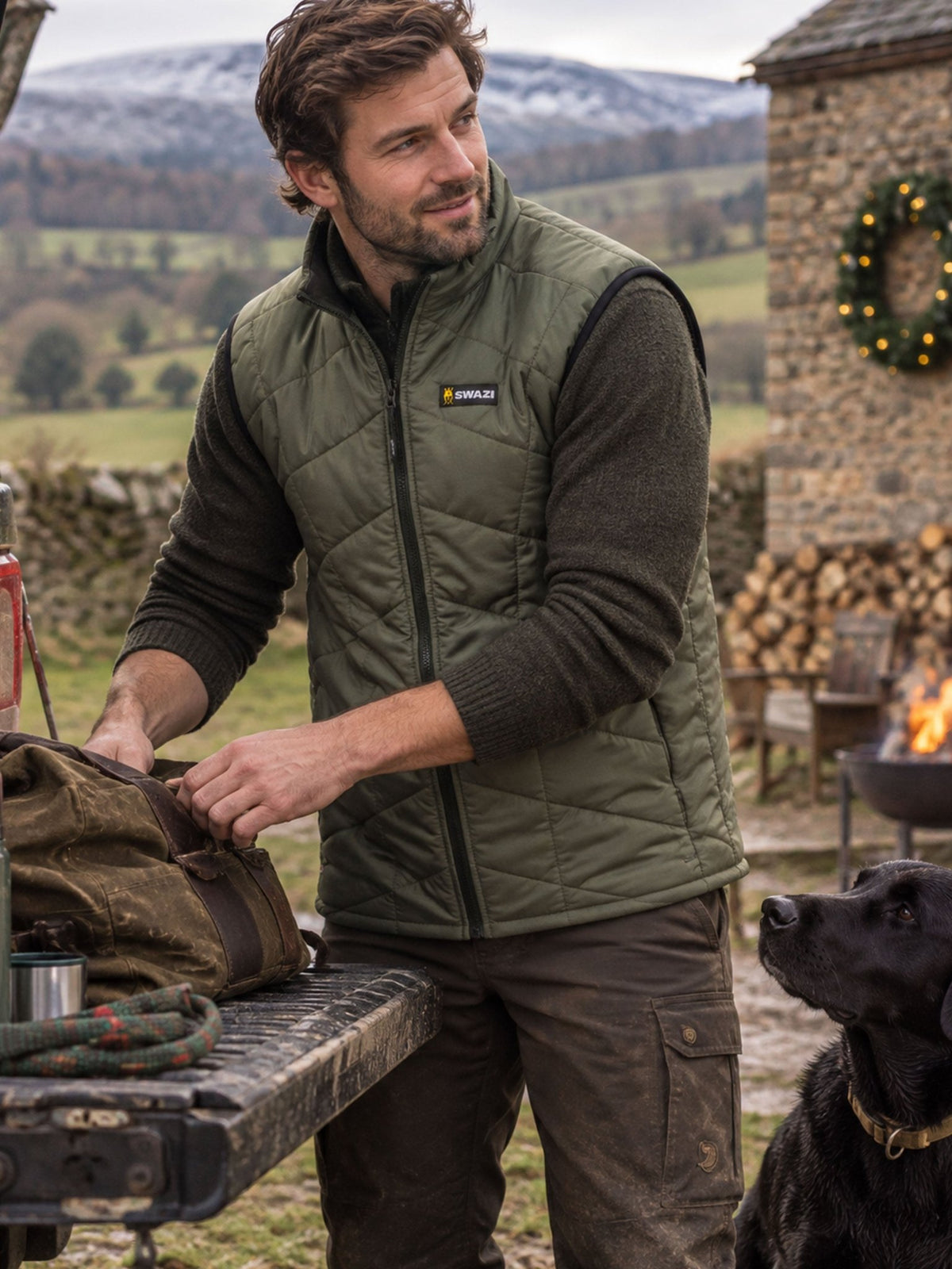 A man wearing an olive green Patagonia zip-up puffer vest over a dark long-sleeve shirt stands outdoors near a stone structure and fire pit, packing a brown bag on a table. A black dog is visible at the lower right, with a mountain landscape in the background.